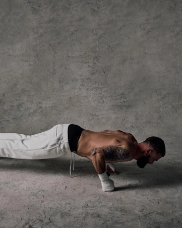 Man doing a one-handed push-up in a studio. He wears white sweatpants and has a tattoo on his arm. Grey concrete background and floor.
