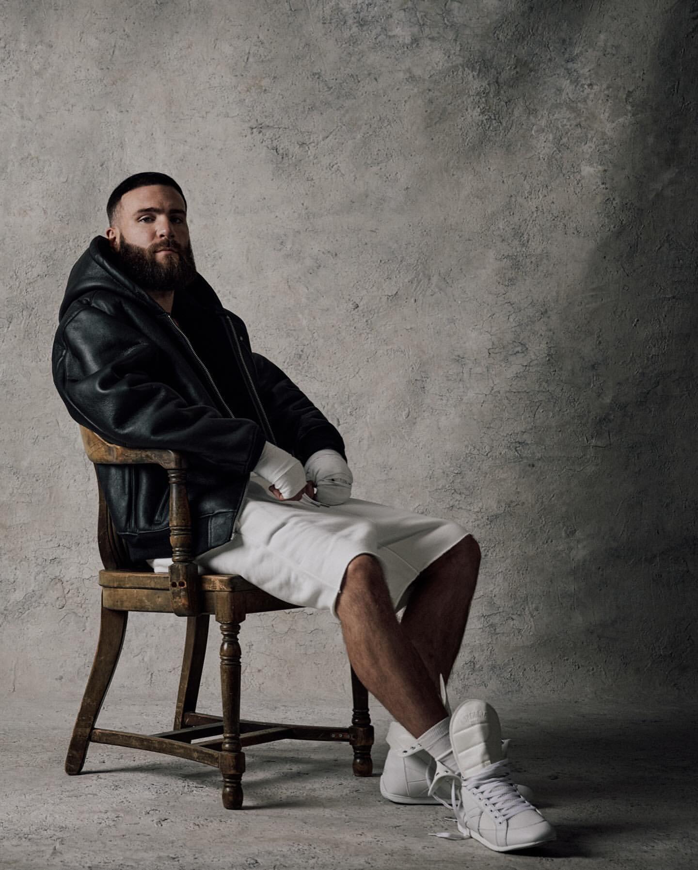 Portrait of a bearded man in a leather jacket sitting on a wooden chair, wearing shorts, and white sneakers in a photo studio.