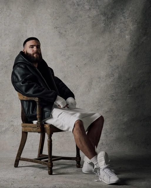 Portrait of a bearded man in a leather jacket sitting on a wooden chair, wearing shorts, and white sneakers in a photo studio.