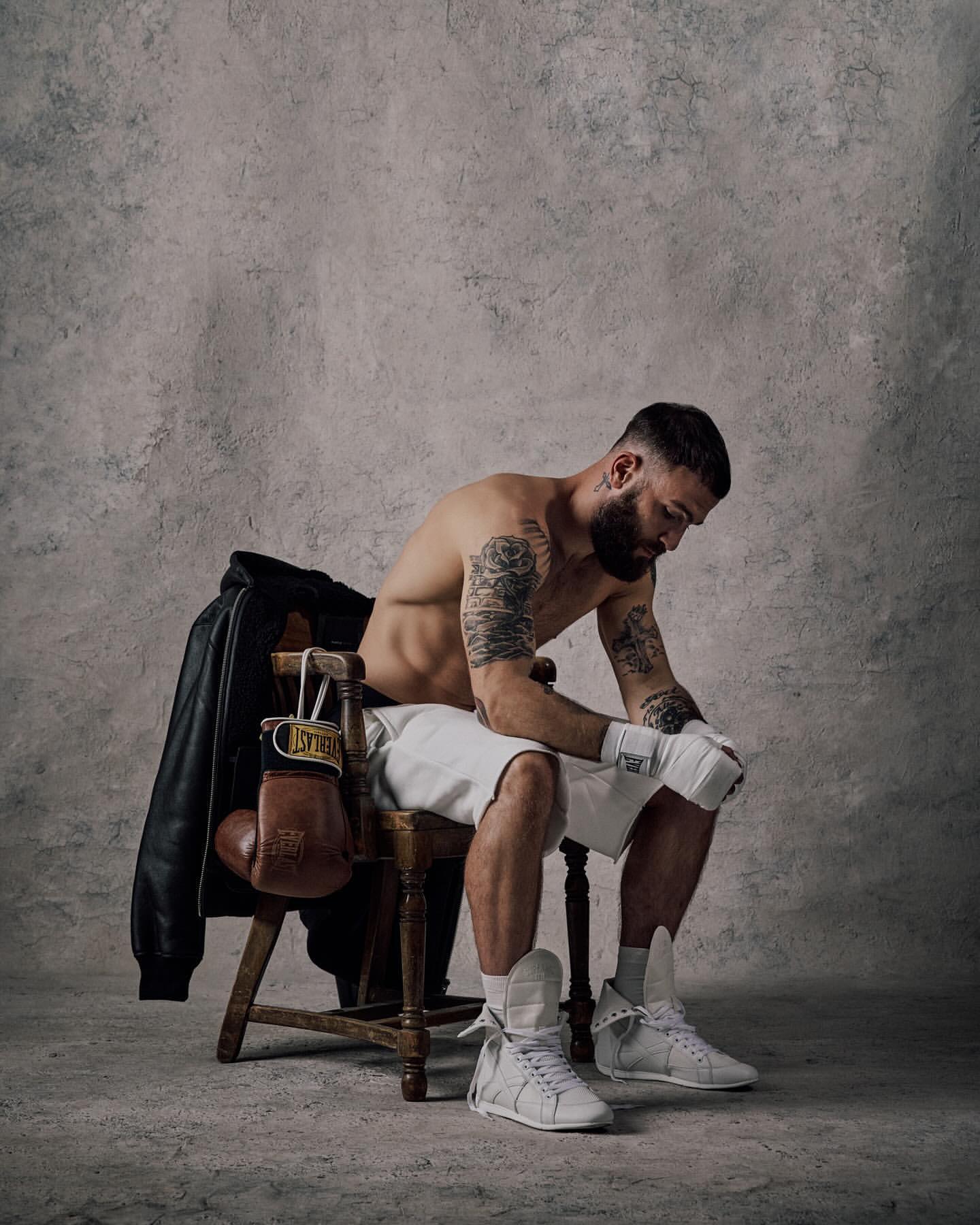 Boxer sits on a chair in a photo studio, wearing boxing gloves and shorts. A leather jacket and boxing gloves hang on the chair.
