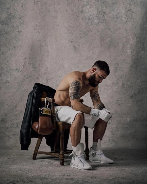 Boxer sits on a chair in a photo studio, wearing boxing gloves and shorts. A leather jacket and boxing gloves hang on the chair.
