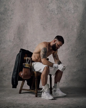 Boxer sits on a chair in a photo studio, wearing boxing gloves and shorts. A leather jacket and boxing gloves hang on the chair.