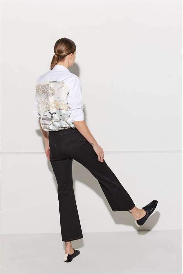 Back view of a stylish adult woman in black pants and a white shirt with a graphic design, striking a dynamic pose against a white backdrop.