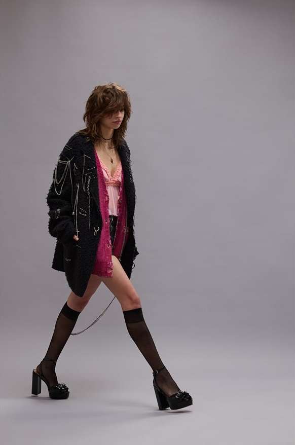 A slender woman strides confidently in a studio, showcasing high-heeled platform shoes, knee-high socks, and a stylishly casual layered outfit.