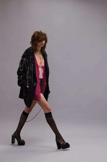 A slender woman strides confidently in a studio, showcasing high-heeled platform shoes, knee-high socks, and a stylishly casual layered outfit.