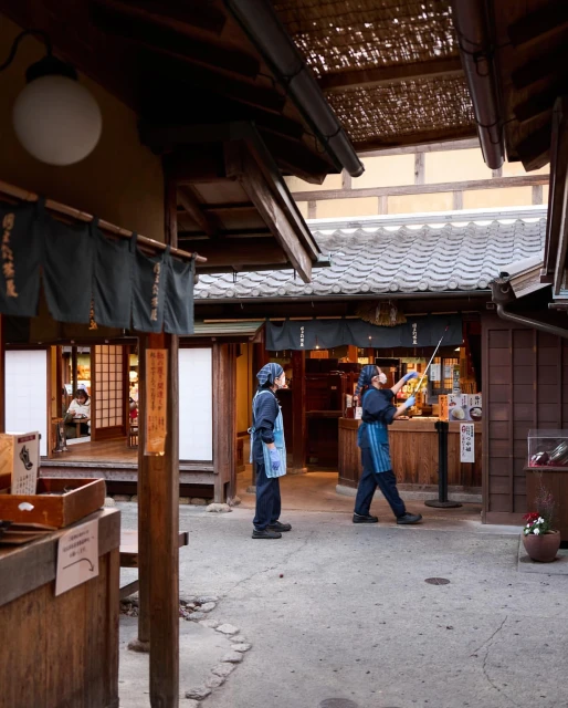 Two individuals wearing aprons and head coverings stand in an outdoor alleyway in front of Japanese-style buildings with dark tile roofs.