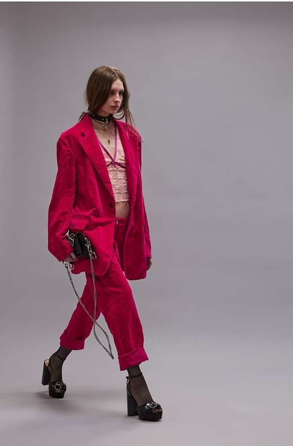 A woman walks wearing a pink suit, bralette, choker necklace, and platform heels. She carries a black purse with a chain.