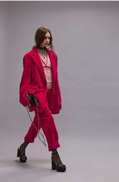 A woman walks wearing a pink suit, bralette, choker necklace, and platform heels. She carries a black purse with a chain.