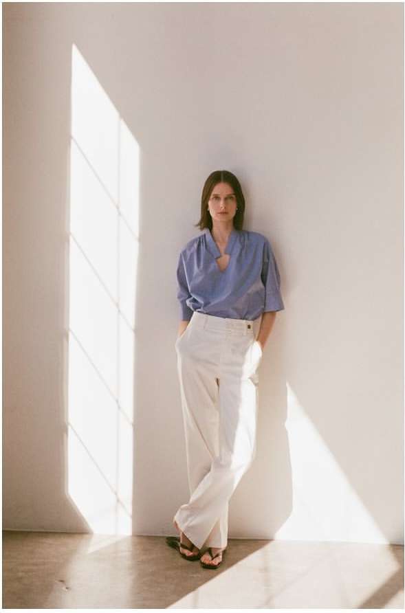 A brunette woman in a blue top and white pants leans against a white wall with natural light creating shadows, in a fashion pose.