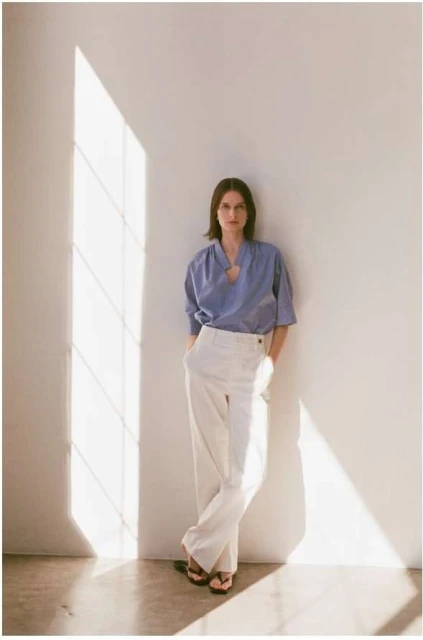 A brunette woman in a blue top and white pants leans against a white wall with natural light creating shadows, in a fashion pose.