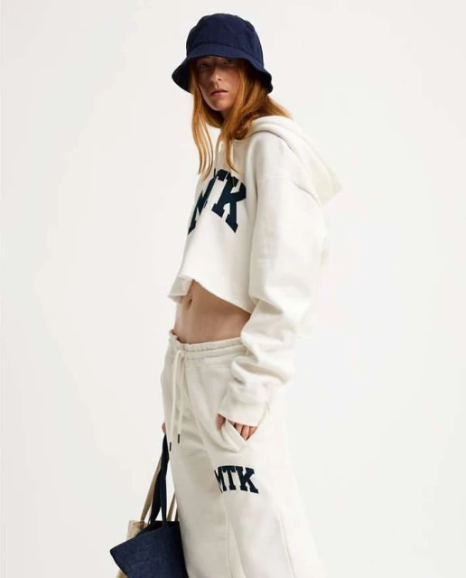 Fashion shot of a woman wearing a white crop top hoodie, sweatpants, and a bucket hat, holding a tote bag against a plain background.