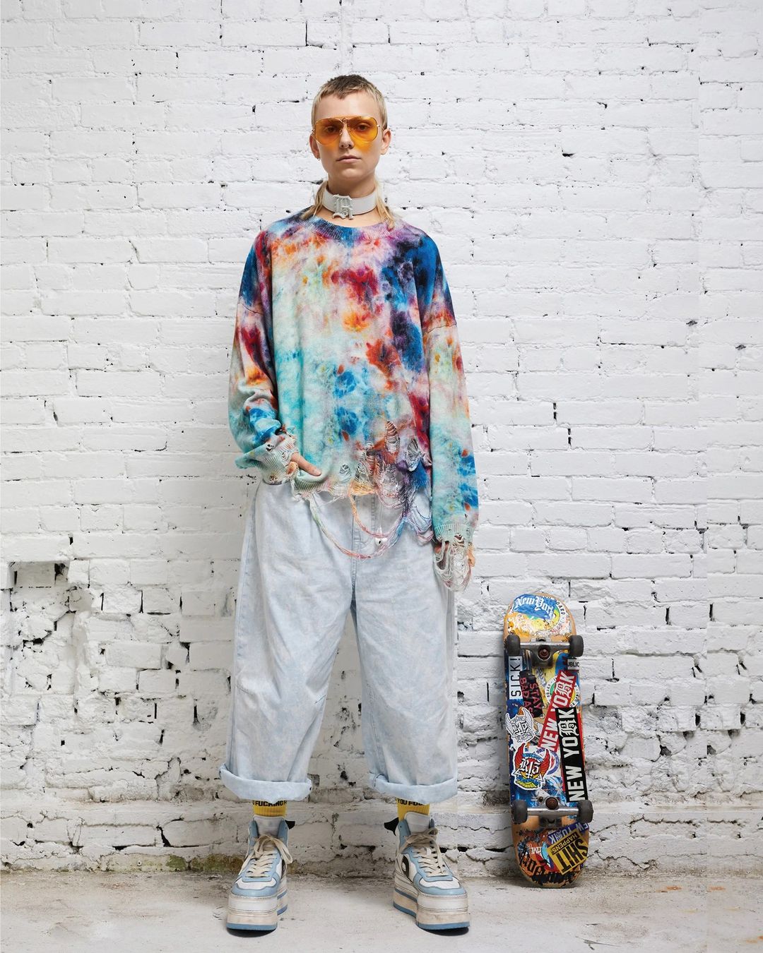Full-length studio shot of a young adult with a tie-dye shirt and platform shoes, standing next to a skateboard against a white brick wall.