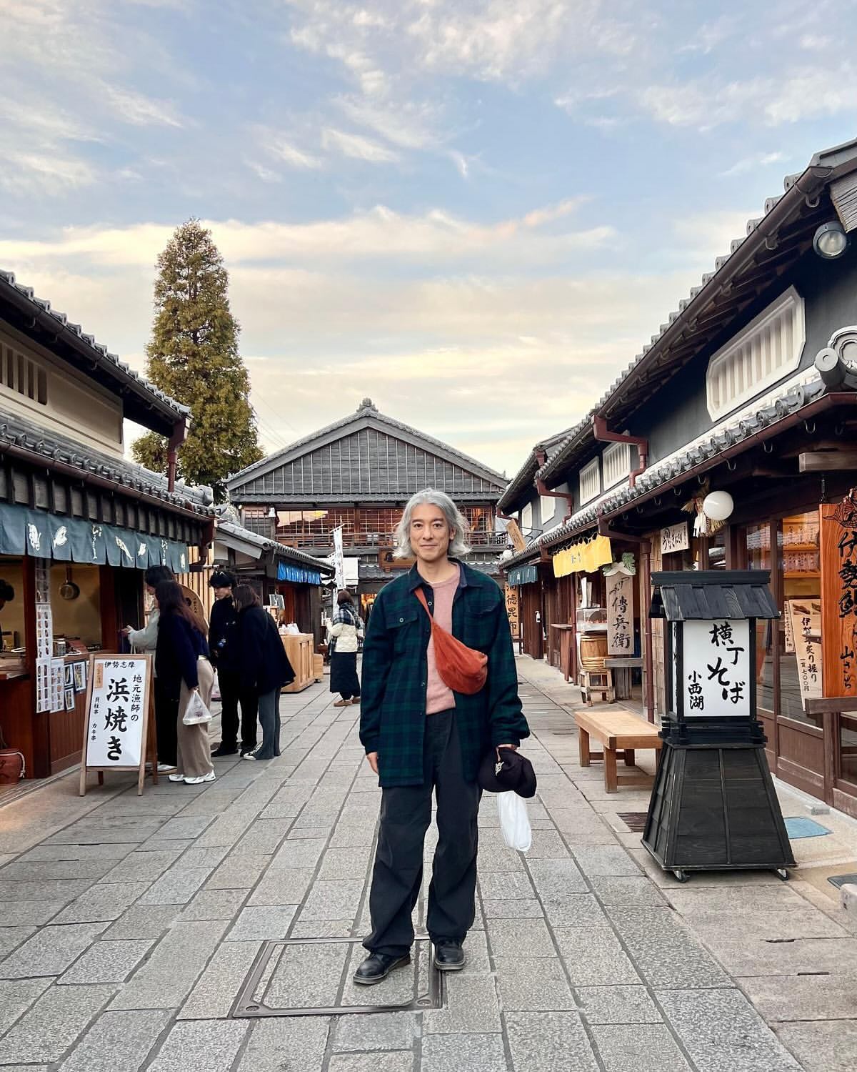 Adult with grey hair stands on cobblestone street in traditional Japanese village with timber buildings and cloudy sky.