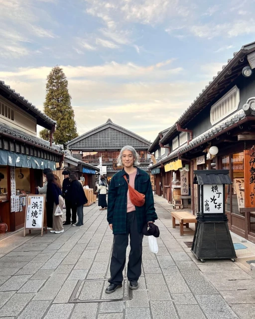 Adult with grey hair stands on cobblestone street in traditional Japanese village with timber buildings and cloudy sky.
