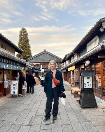 Adult with grey hair stands on cobblestone street in traditional Japanese village with timber buildings and cloudy sky.