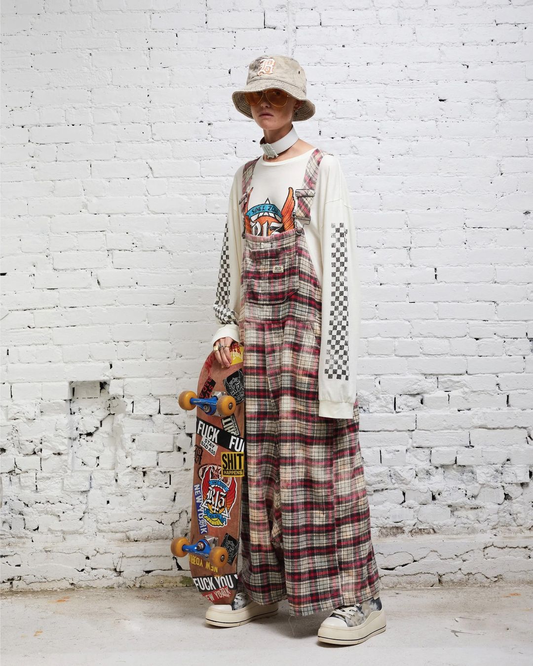 A person in overalls and a bucket hat poses against a white brick wall, holding a sticker-covered skateboard in a studio shot.