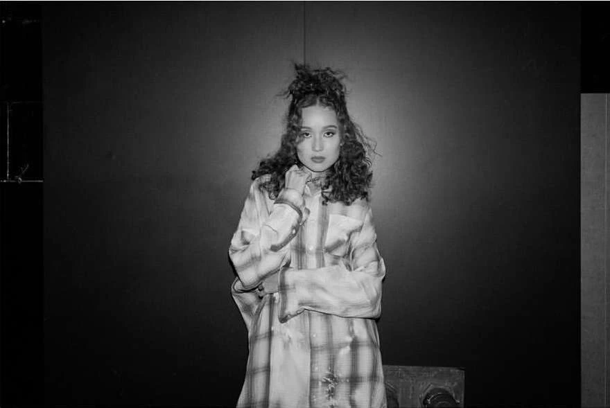 Black and white portrait of a young woman with curly hair wearing a plaid shirt standing against a dark studio backdrop.