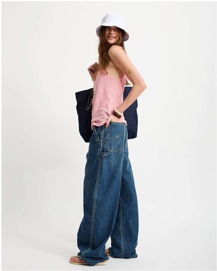 Young woman in casual outfit poses with tote bag in a studio setting, wearing pink top, oversized jeans, and bucket hat against a white background.