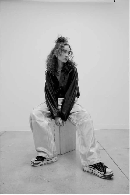 A young adult woman with curly hair sits on a box in an indoor photo studio, wearing a black jacket and oversized denim pants, in black and white.