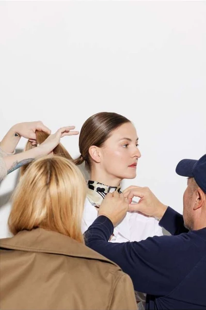 Model getting ready for photoshoot with hair and make up artists assisting. Woman wears white shirt and scarf, in photo studio.