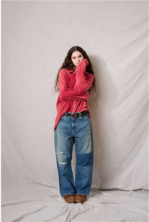 A young adult female model poses in a red collared shirt, jeans, and loafers in a studio with a neutral backdrop, looking at the camera.