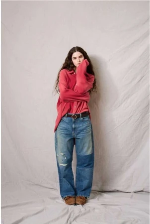 A young adult female model poses in a red collared shirt, jeans, and loafers in a studio with a neutral backdrop, looking at the camera.
