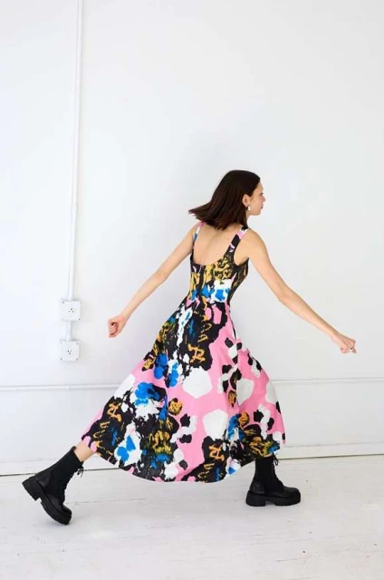 A woman strikes a dynamic pose in a colorful patterned dress and chunky black boots against a stark white studio backdrop.
