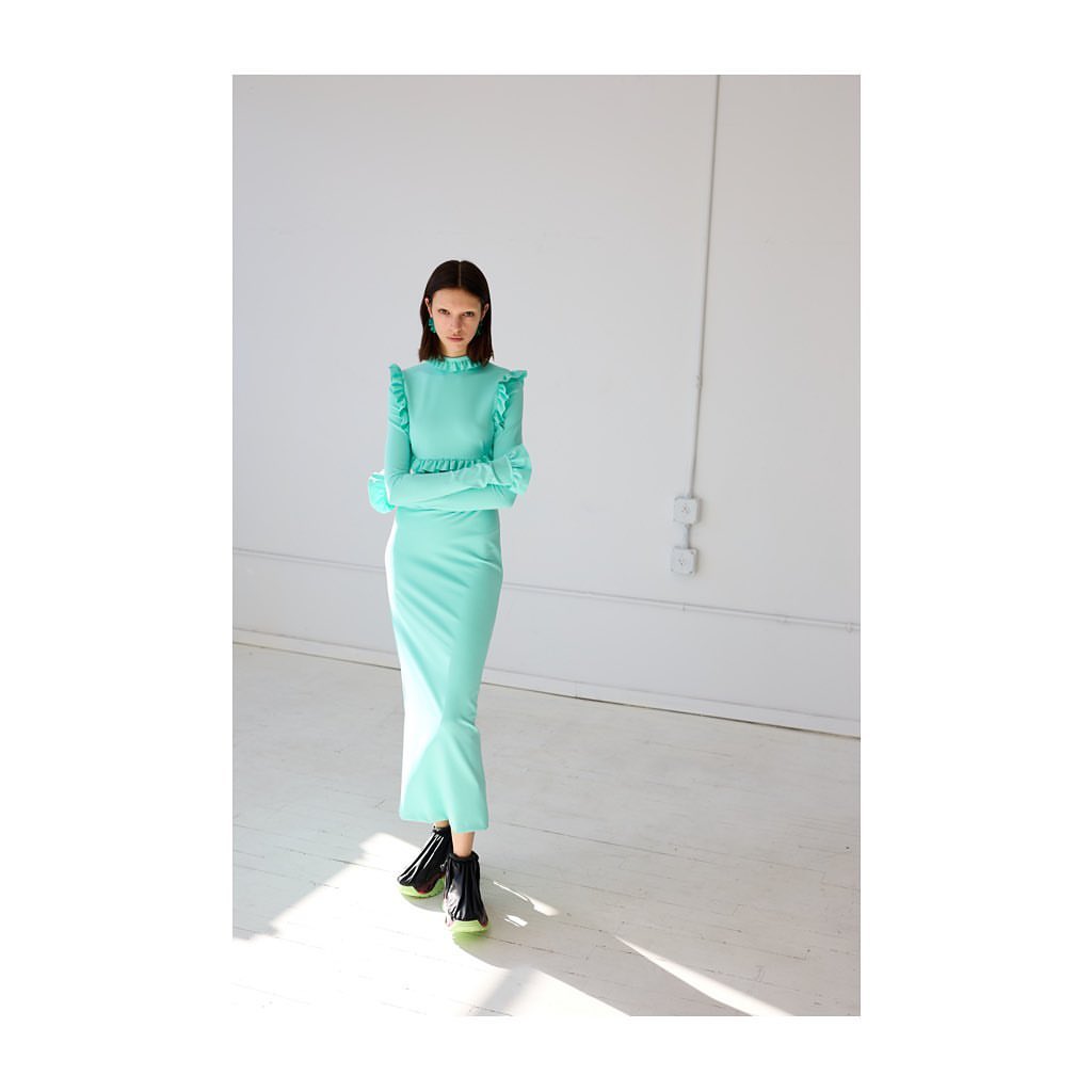 An adult female fashion model poses in a long-sleeved, light-green dress and black ankle boots in a bright white photo studio.