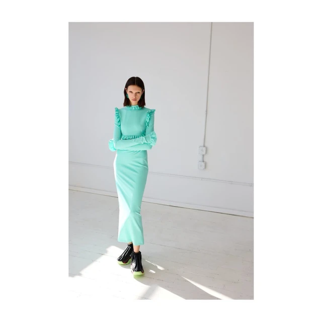 An adult female fashion model poses in a long-sleeved, light-green dress and black ankle boots in a bright white photo studio.