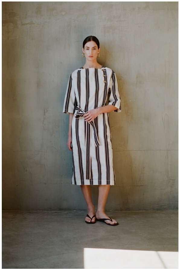 Woman wearing a belted striped dress and sandals standing against a textured concrete wall by Masato Onoda