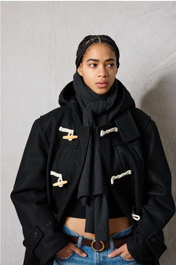 A young adult woman with braided hair poses indoors in a hooded black duffel coat, gray scarf, blue jeans, and brown leather belt.