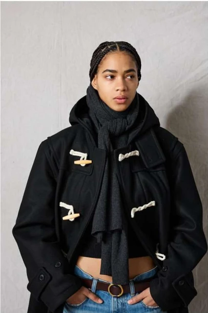 A young adult woman with braided hair poses indoors in a hooded black duffel coat, gray scarf, blue jeans, and brown leather belt.