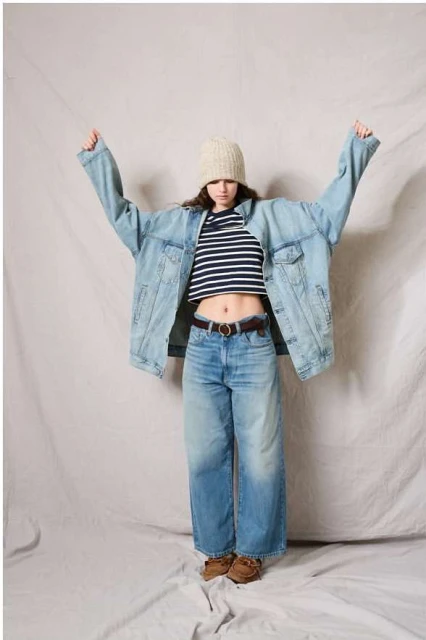 A young woman poses in an oversized light-blue denim jacket, cropped striped top, wide-leg jeans, knit hat, and brown shoes on a grey background.