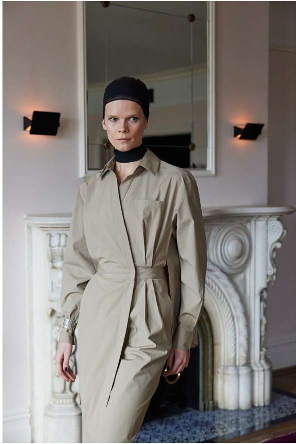Adult female model in a beige shirt dress and black cap poses indoors in front of a fireplace and mirror, fashion photography.