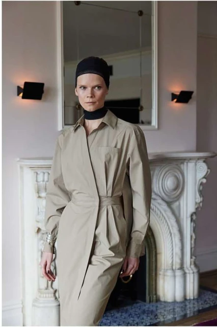 Adult female model in a beige shirt dress and black cap poses indoors in front of a fireplace and mirror, fashion photography.