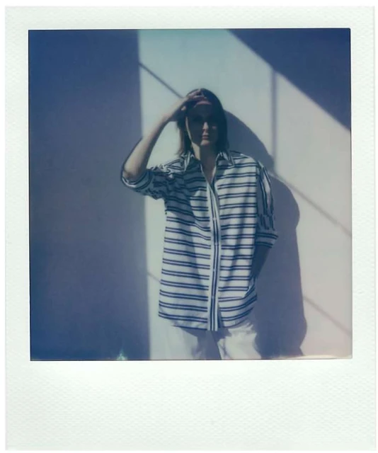 An adult person wearing a striped shirt poses against a wall with sunlight, holding their hand to their forehead in a polaroid style.