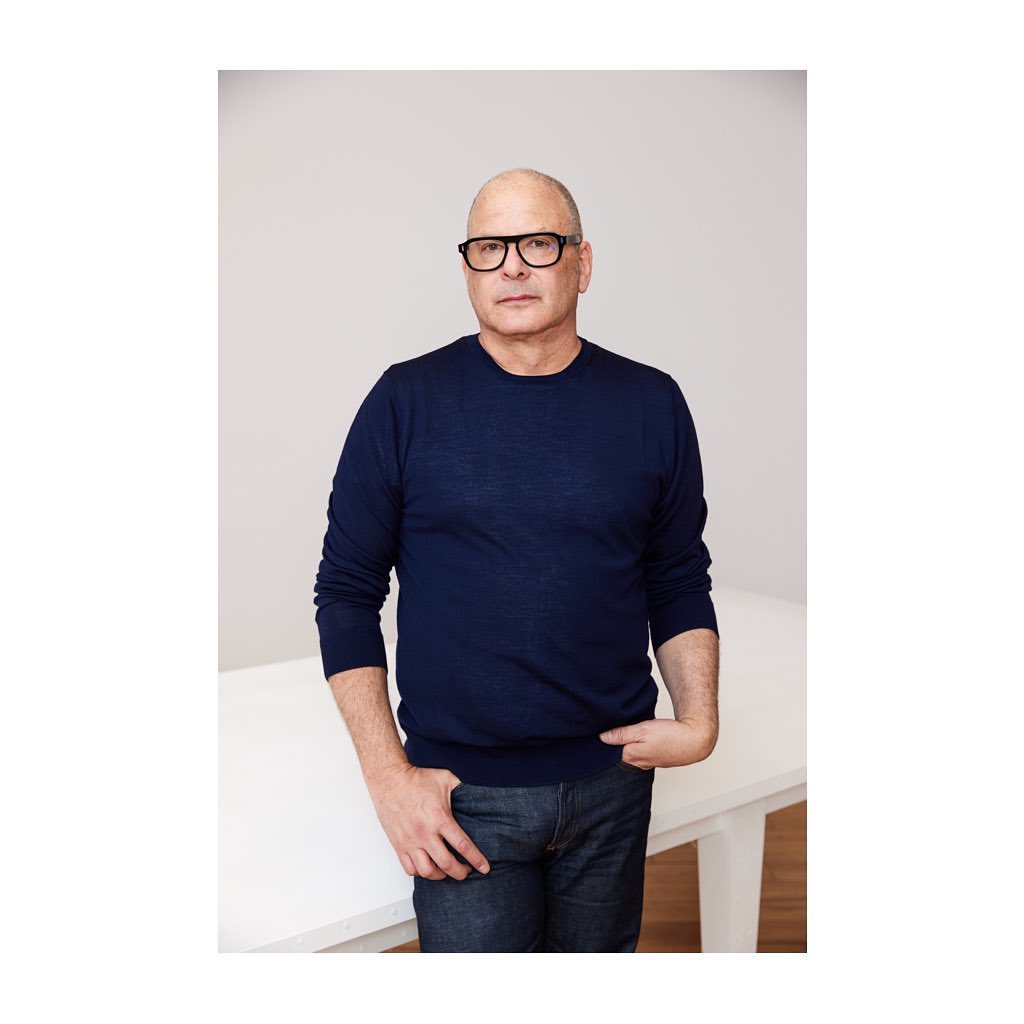 Bald man wearing black glasses and a blue long-sleeve sweater posing against a white studio background.