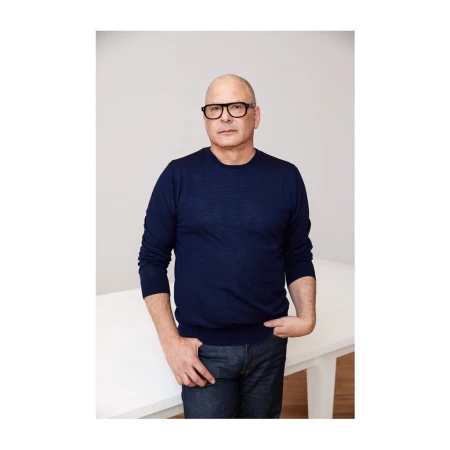 Bald man wearing black glasses and a blue long-sleeve sweater posing against a white studio background.