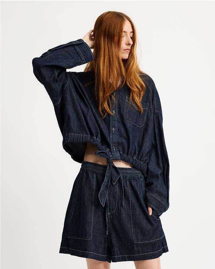 Woman with long red hair wearing a dark denim button-down shirt and matching shorts against a white backdrop