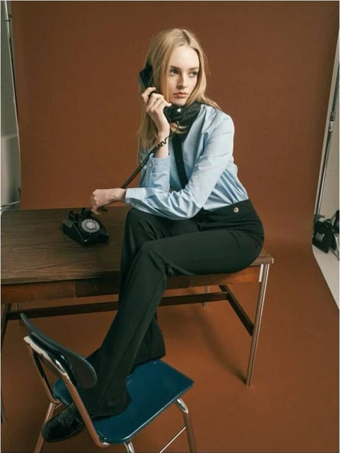 A fair-skinned woman with blonde hair is sitting at a desk while talking on a vintage telephone in a photo studio with brown backdrop.