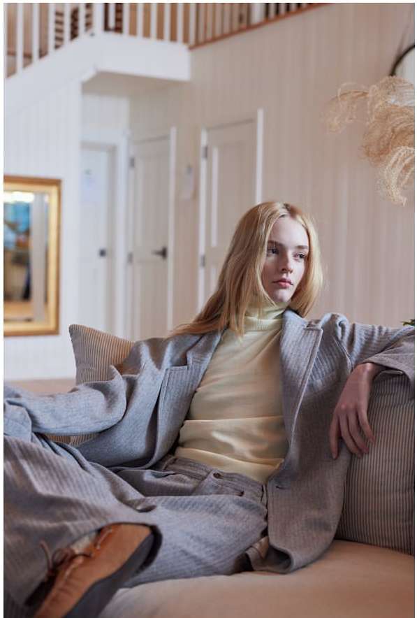 Stylish blonde model lounges in a gray suit on a neutral couch in a bright, naturally lit interior with neutral color palette and modern decor.