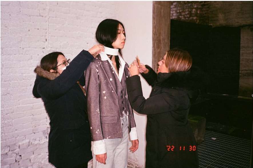 Fashion shoot backstage featuring a model getting adjusted by two stylists, set against a textured brick wall backdrop.