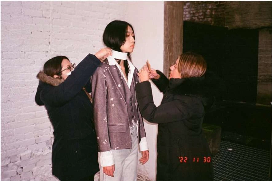 Fashion shoot backstage featuring a model getting adjusted by two stylists, set against a textured brick wall backdrop.
