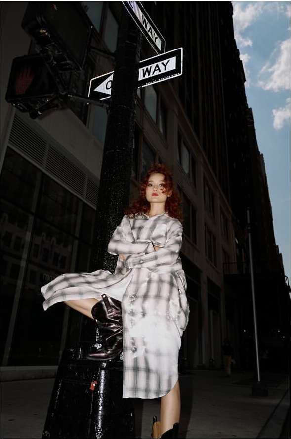 Young woman with auburn hair posing on a street post in an oversized plaid shirt dress and black boots, buildings in background.