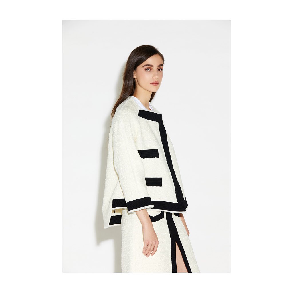 A brunette fashion model poses in a cream-colored bouclé jacket with black trim and a matching skirt against a white backdrop.