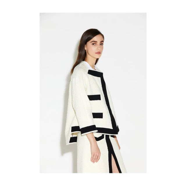 A brunette fashion model poses in a cream-colored bouclé jacket with black trim and a matching skirt against a white backdrop.