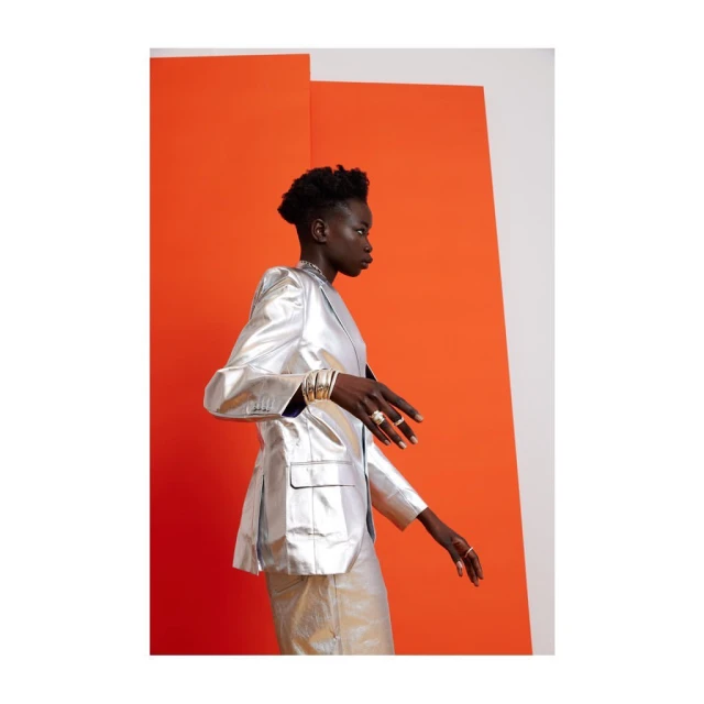 A striking portrait of an adult female model in a silver suit against an orange and white background, showcasing fashion and style.