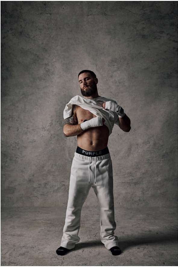 Man in sweatpants and a t-shirt stands in front of a gray textured backdrop in a studio setting, exhibiting a casual and relaxed pose.
