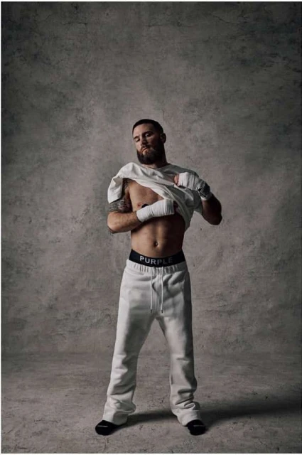 Man in sweatpants and a t-shirt stands in front of a gray textured backdrop in a studio setting, exhibiting a casual and relaxed pose.