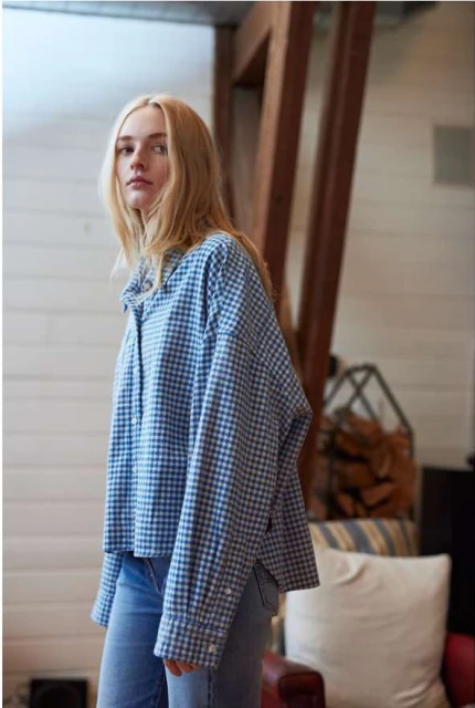Adult female poses indoors wearing a blue and white gingham button-up shirt and blue jeans, illuminated by natural light.